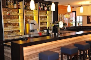 Una zona de bar con estanterías llenas de copas y botellas, una barra y taburetes azules.