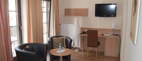 A hotel room with a bed, two armchairs, a small round table with a bottle, a desk with a chair, a flat-screen TV, and a radiator.