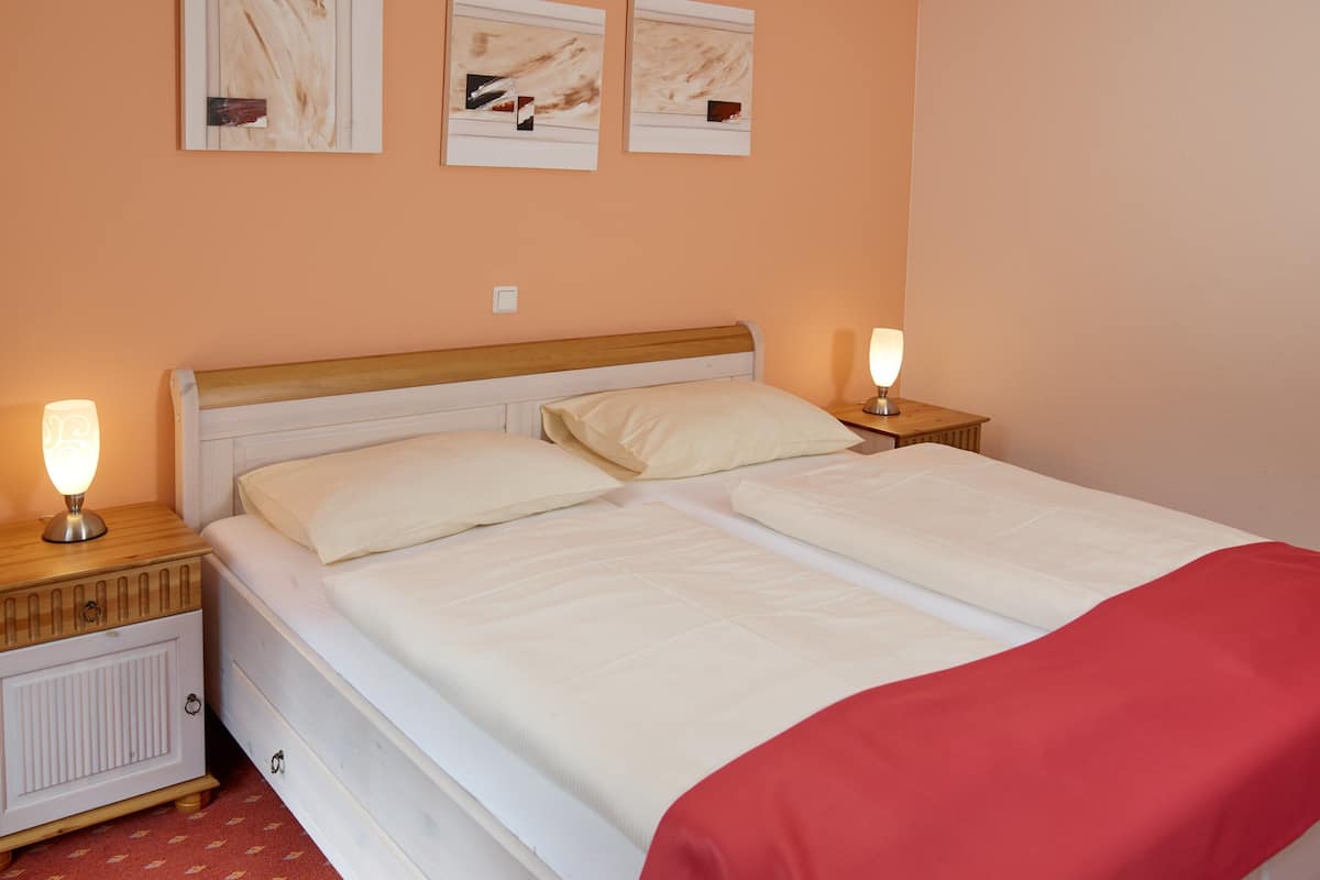A double bed with white bedding, two bedside tables with lamps, a wooden headboard, and a red blanket on the right side.