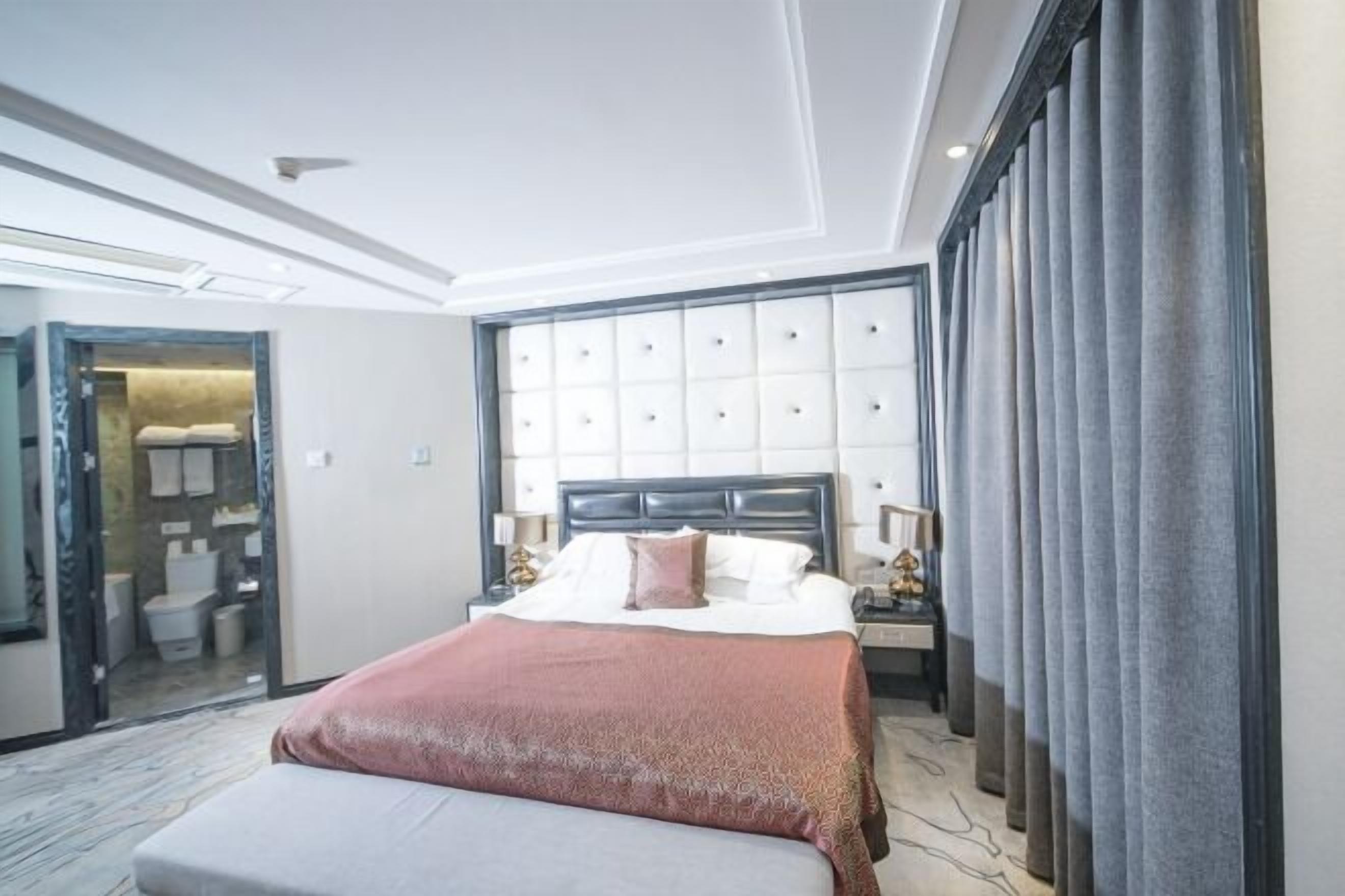 Una habitación de hotel con una cama grande, una mesita de noche con lámpara, un armario y un baño visible a través de una puerta abierta.