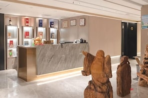 A reception desk with a marble counter, a reception area with shelves displaying items, and two wooden sculptures. - Victoria Royal Hotel (Shangrao)