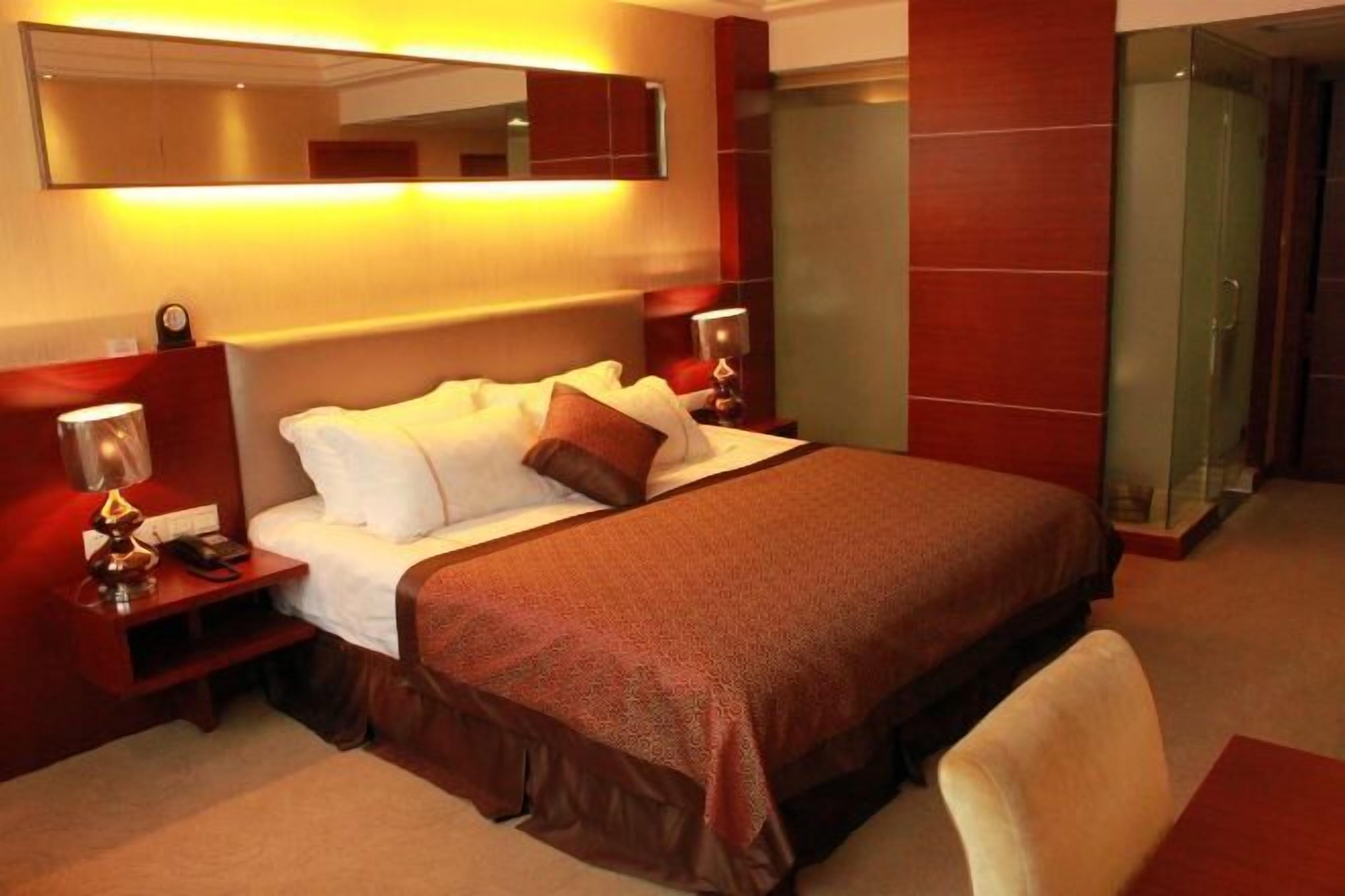 A hotel room with a large bed, a bedside table with a lamp, a mirror above the bed, and a glass-enclosed shower area.