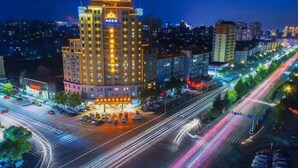A multi-story hotel with a prominent illuminated facade, a parking lot, and a busy urban street with light trails from moving vehicles. - Victoria Royal Hotel (Shangrao)