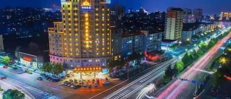 A multi-story hotel with a prominent illuminated facade, a parking lot, and a busy urban street with light trails from moving vehicles.