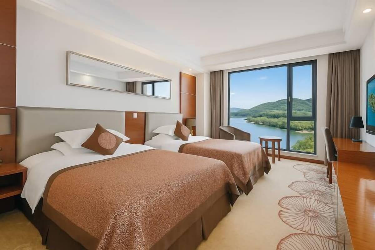 A hotel room with two beds, a large window with a view of a lake and hills, a desk with a chair, and a TV.