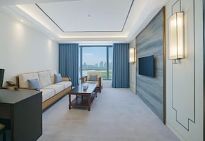 A modern hotel room with a sofa, armchairs, a coffee table, a TV, and a city view. - Victoria Royal Hotel (Shangrao)
