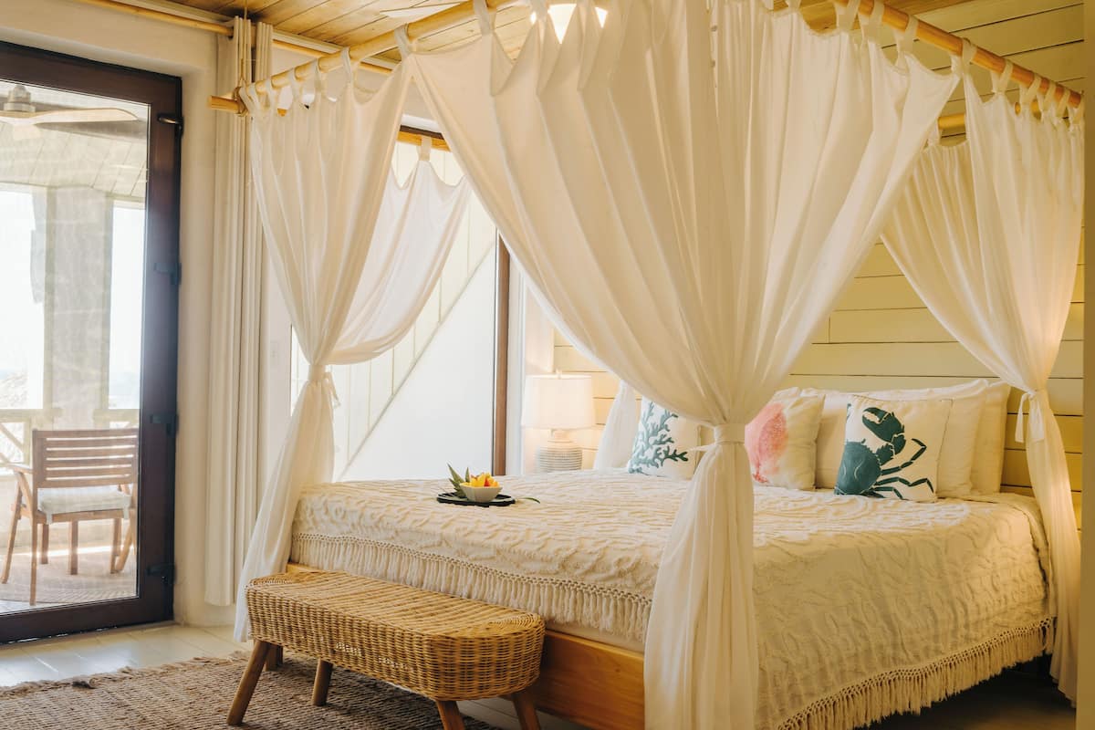 A bedroom with a canopy bed, a wicker bench, and a view of the outdoors.