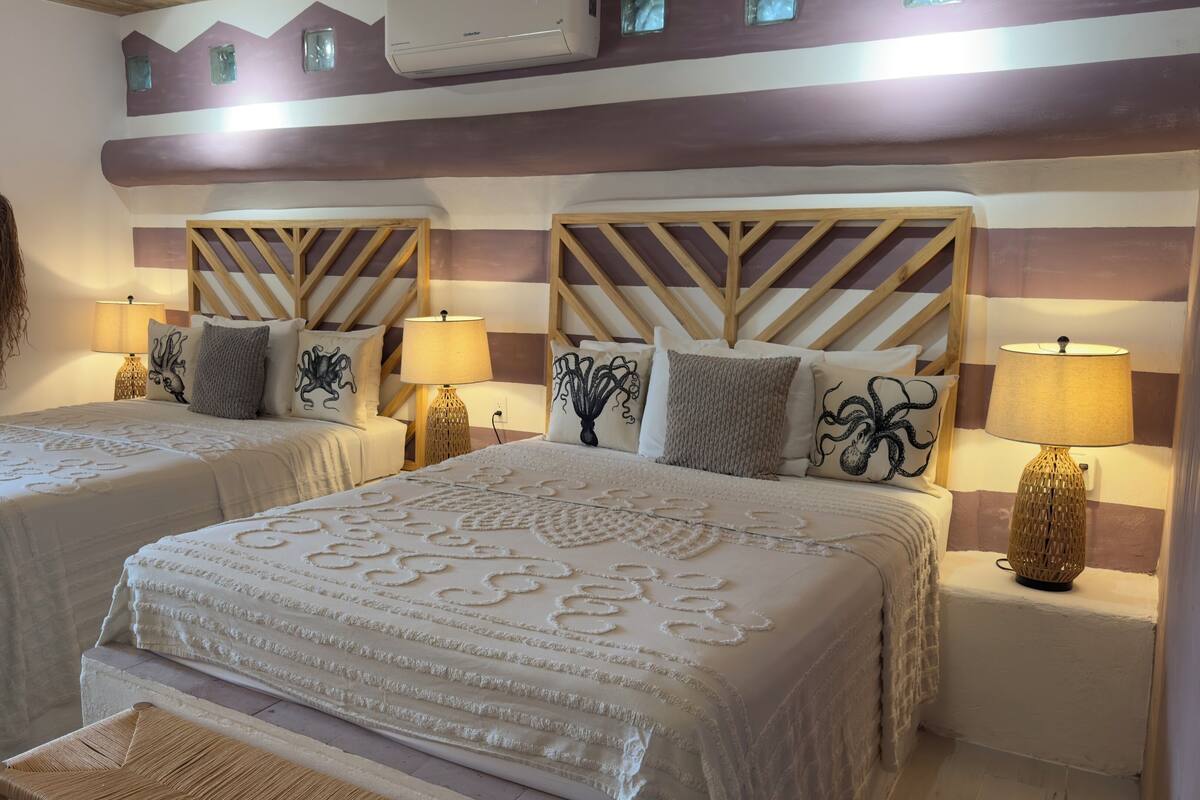 A hotel room with two beds, each with a wooden headboard, white bedding, and decorative pillows. There are two bedside tables with lamps, a wall-mounted air conditioner, and a woven wall hanging.