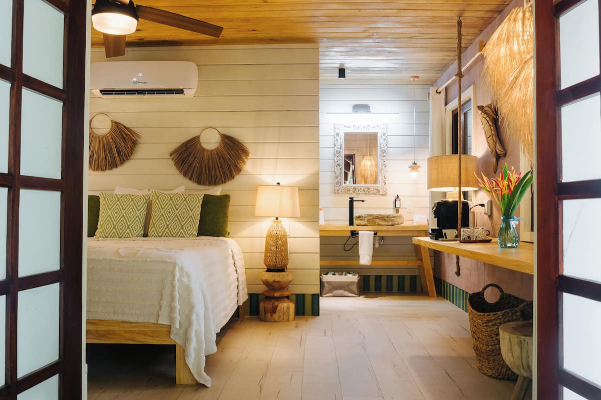 A room with a bed, a bedside lamp, a mirror, a hanging basket, and a small table with a vase.