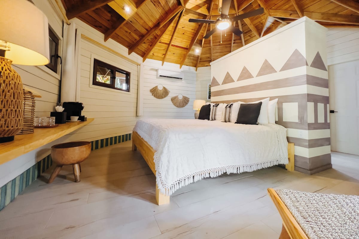 A bedroom with a wooden ceiling, a bed with white bedding, a bedside table, and a wall-mounted air conditioner.