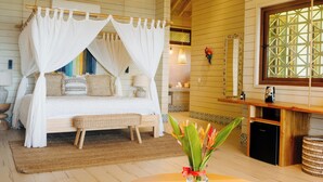 A bedroom with a four-poster bed, a bedside table with a lamp, a small refrigerator, and a window with a wooden frame. - Tranquilseas Boutique Eco Resort & Dive Center (Roatan)