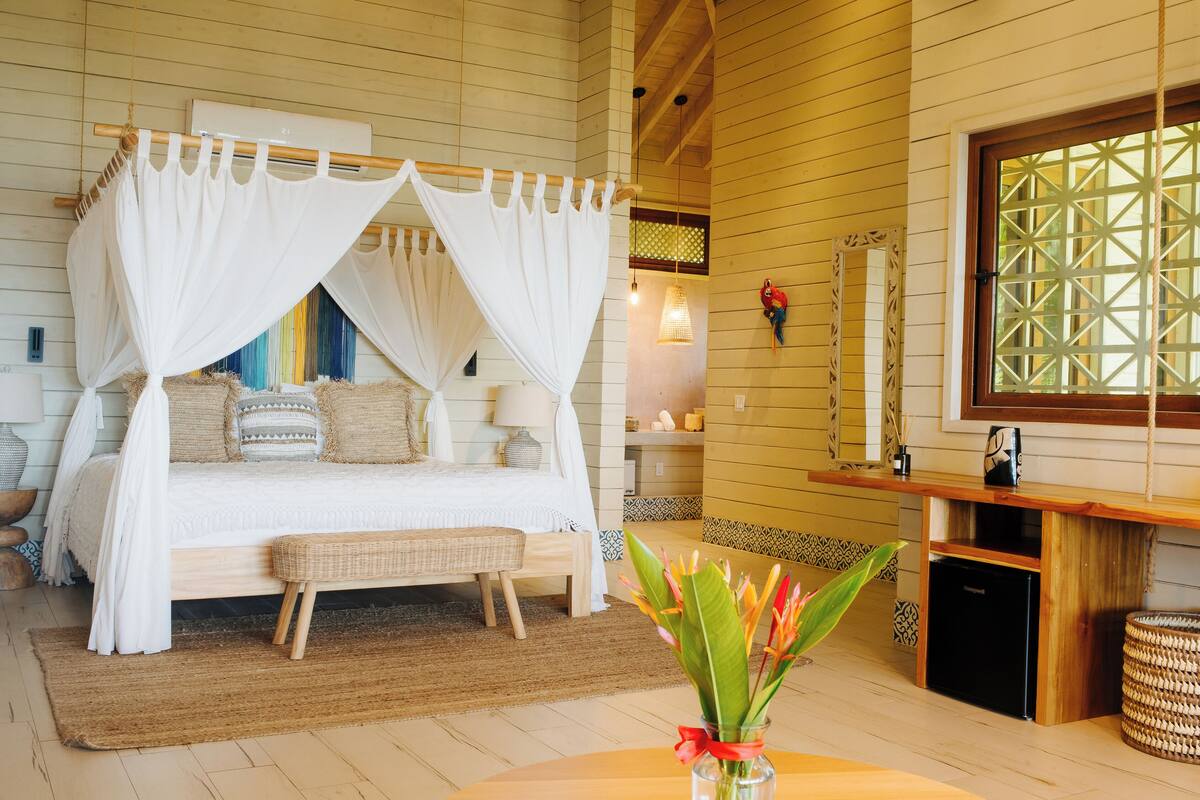 A bedroom with a four-poster bed, a bedside table with a lamp, a small refrigerator, and a window with a wooden frame.