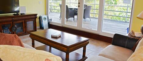 A living room with a sofa, coffee table, and a television. There is a sliding glass door leading to an outdoor patio with chairs and a table.