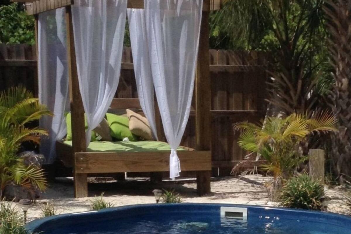 A wooden canopy bed with white curtains, a green cushion, and a small pool in front.