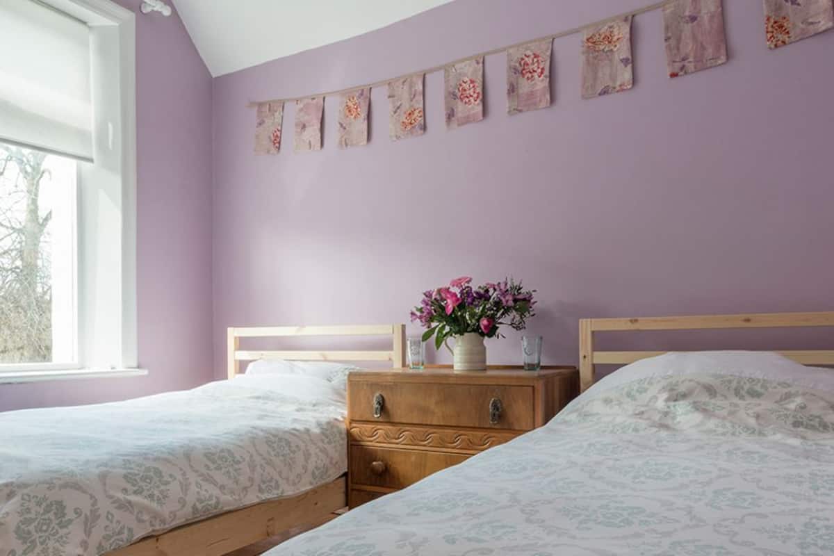 A bedroom with two single beds, a wooden dresser, a nightstand with a vase of flowers, and a window with blinds.