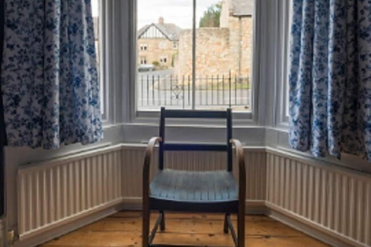 A corner room with a bay window, a wooden chair, and a view of a stone building outside.