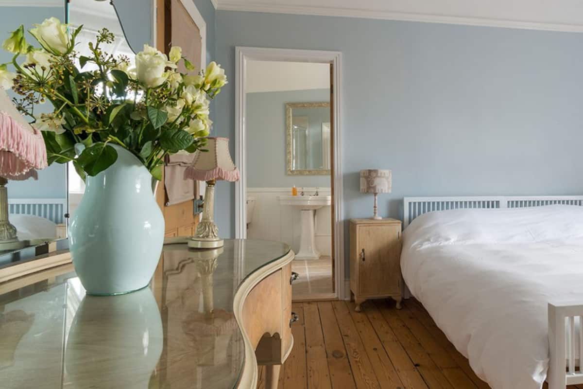 A bedroom with a large bed, a dresser, a vanity with a mirror, and a vase of flowers.