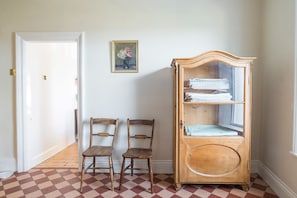 Una habitación con piso a cuadros, dos sillas de madera, un aparador de madera con puertas de cristal y un cuadro en la pared.