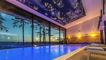 Ein moderner, überdachter Swimmingpool mit Sternenhimmel und Blick auf den Sonnenuntergang durch große Fenster.