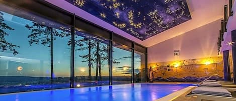 A modern indoor swimming pool with a starry ceiling and a view of the sunset through large windows.