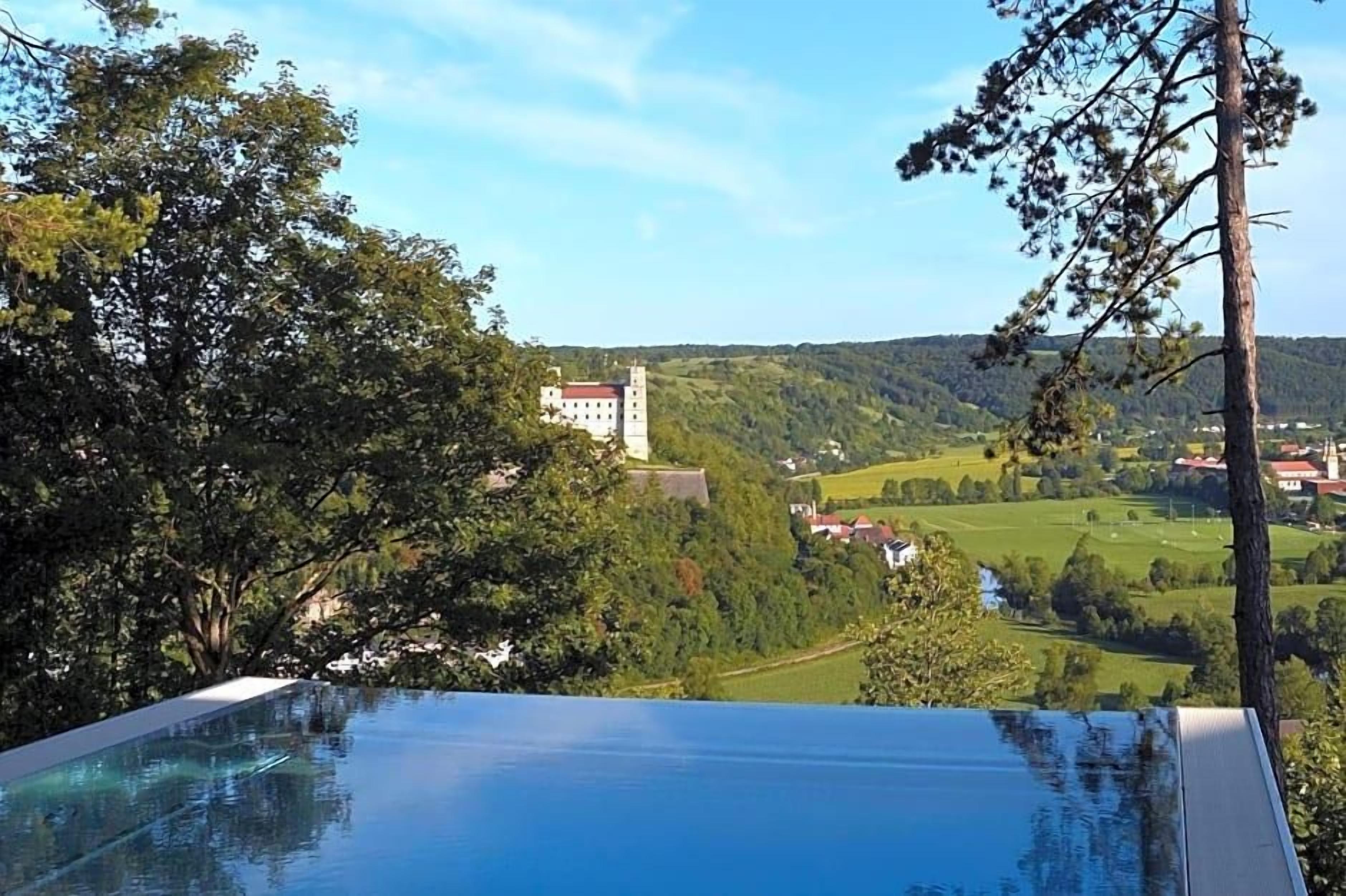 A view of a hotel with a pool overlooking a valley, a building in the distance, and a forested area.