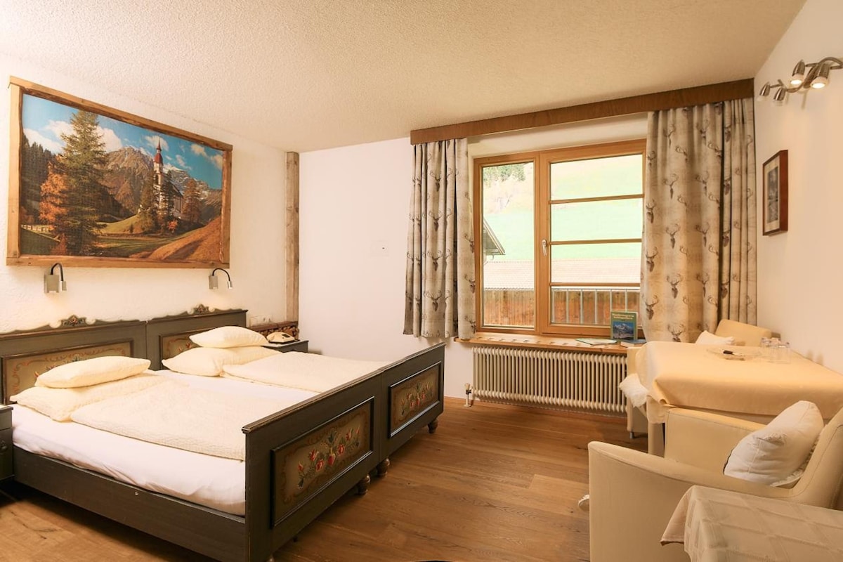 A hotel room with a bed, a chair, a television, a window with curtains, and a painting on the wall.