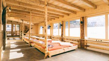 Eine Holzhütte mit mehreren reihenweise angeordneten Betten, großen Fenstern und einem steinbelagten Boden.