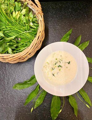 Un tazón blanco de sopa con hierbas verdes sobre una superficie oscura, acompañado de una cesta de mimbre con hojas verdes.