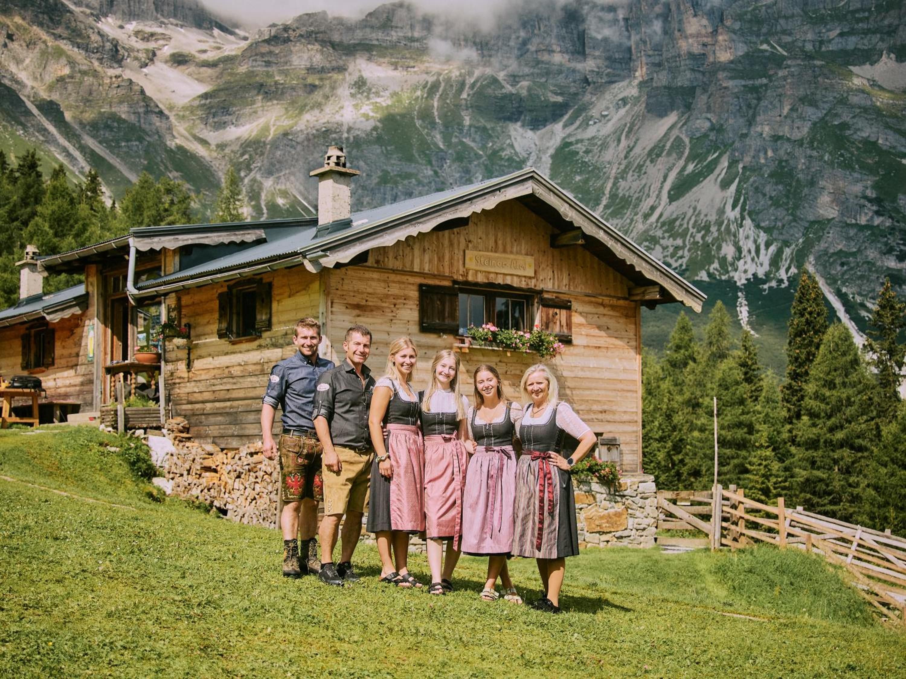 Eine Holzhütte mit Steinunterbau, umgeben von einem Holzzaun, vor einer Bergkulisse gelegen.