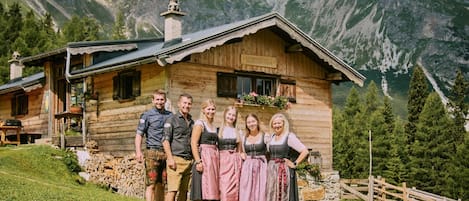 A wooden cabin with a stone foundation, surrounded by a wooden fence, set against a mountainous backdrop.