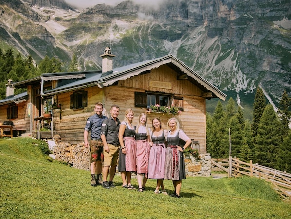 A wooden cabin with a stone foundation, surrounded by a wooden fence, set against a mountainous backdrop.
