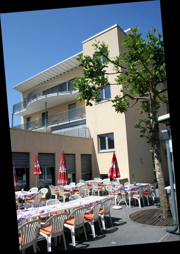An outdoor dining area with tables and chairs, red umbrellas, and a building with a balcony.