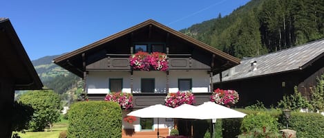 A traditional chalet with a balcony adorned with flowers, outdoor seating, and a view of a mountainous landscape.
