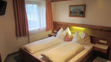 A double bed with white linens and pillows, a wooden headboard, a painting on the wall, a window with curtains, and a small bedside table.