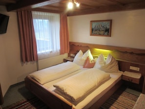 A double bed with white linens and pillows, a wooden headboard, a painting on the wall, a window with curtains, and a small bedside table.