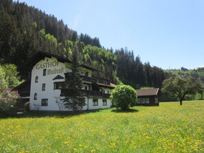 A building labeled 'GASTHOF Waldgraft' with a traditional architectural style, surrounded by a lush green field and a forested hillside.