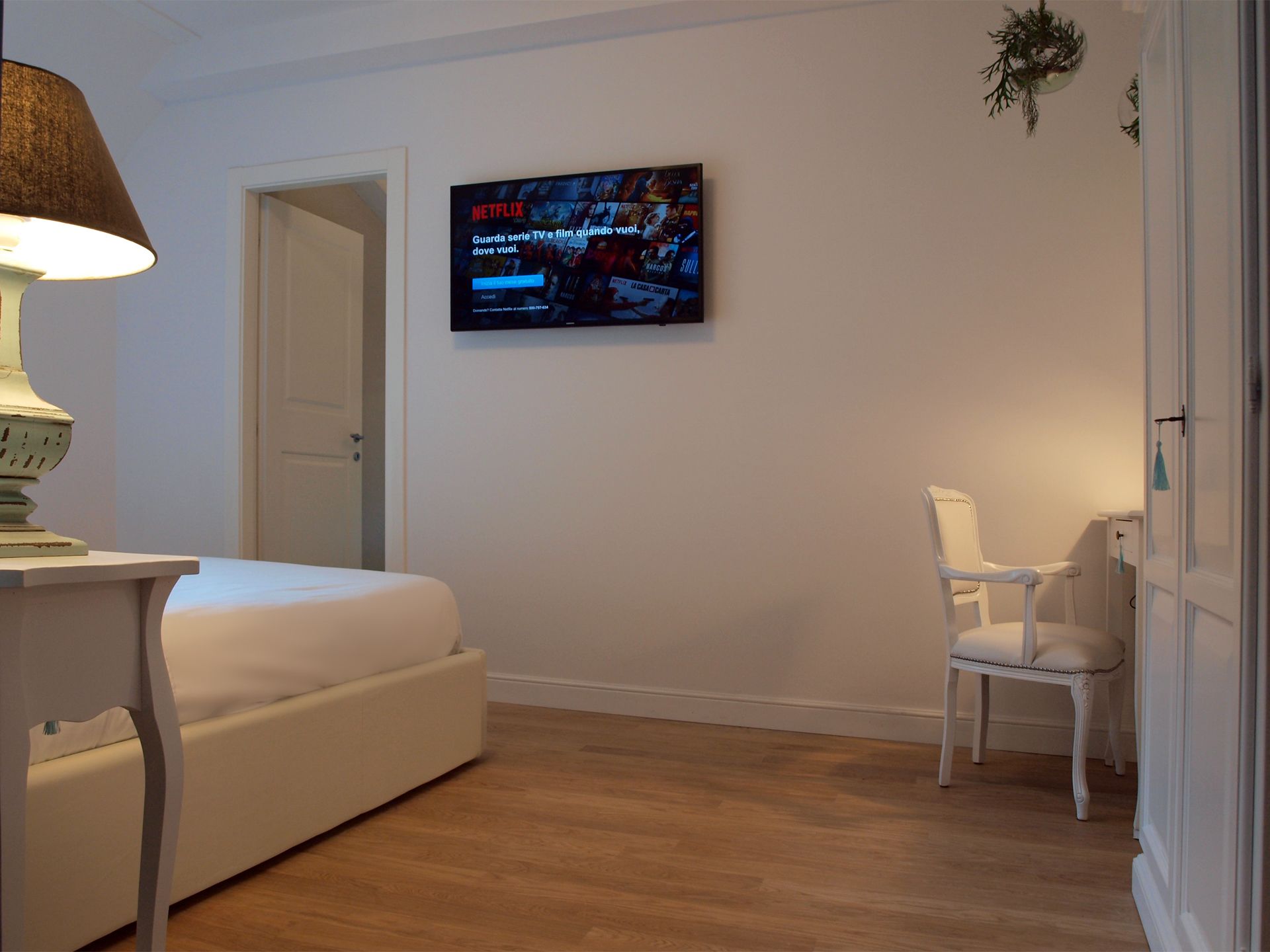 Una habitación con una cama, una silla, un televisor montado en la pared y una lámpara sobre una mesa.