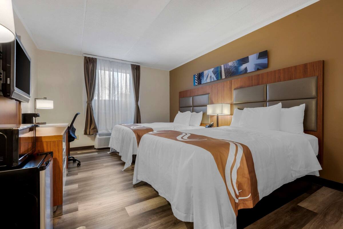 A hotel room with two beds, a desk with a microwave, a TV, and a large window with curtains.