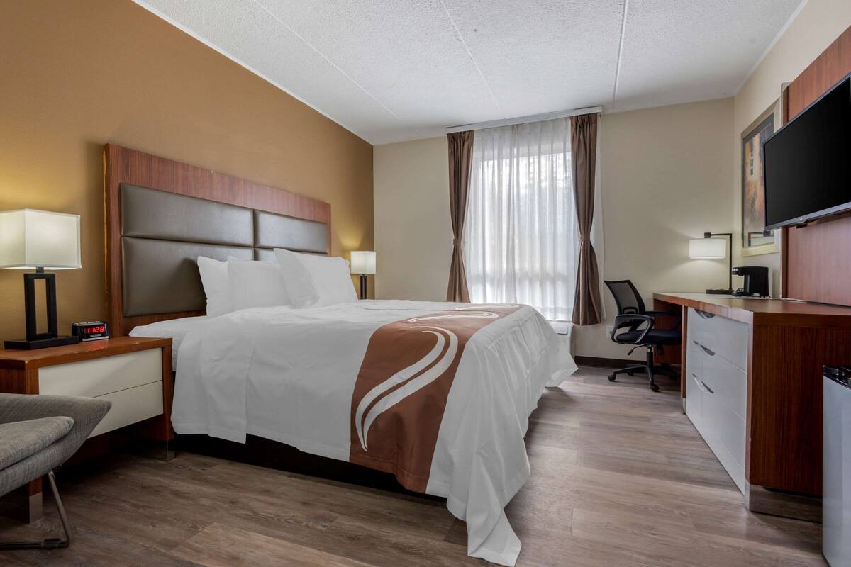A hotel room with a bed, a desk, a chair, a TV, and a window with curtains.