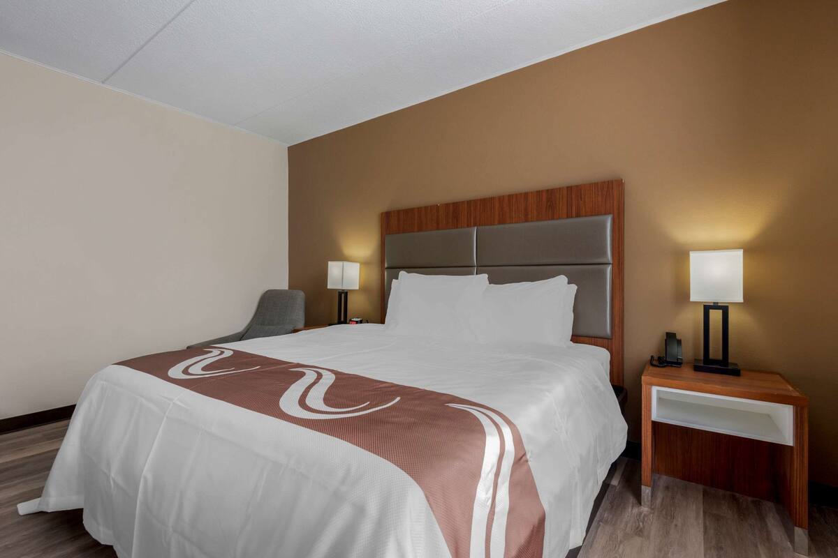 A hotel room with a large bed, two bedside tables with lamps, a chair, and a wooden headboard.