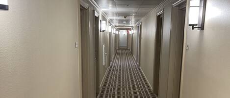 Un couloir d’hôtel avec un tapis à motifs, des portes et des luminaires fixés au mur.