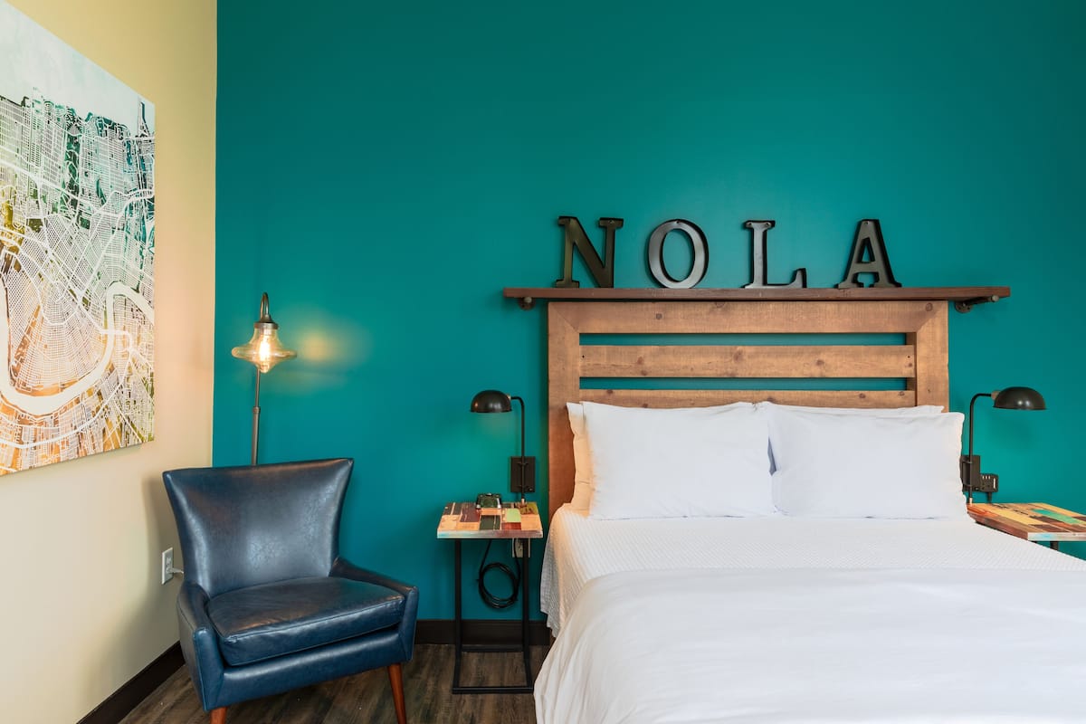 Une chambre d’hôtel avec un mur de couleur turquoise, une tête de lit en bois ornée des lettres « NOLA », un lit avec du linge de lit blanc, un fauteuil en cuir bleu et une petite table avec une lampe.