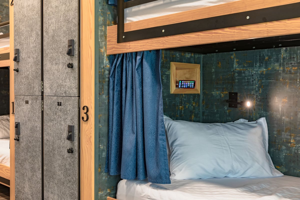 Un lit superposé avec une tête de lit en bois, un lit simple avec un matelas blanc, un rideau bleu et une petite horloge numérique accrochée au mur.