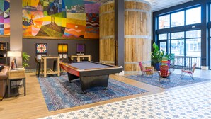 A pool table in a room with a colorful mural, wooden accents, and a view through large windows. - HI New Orleans - Hostel (New Orleans)