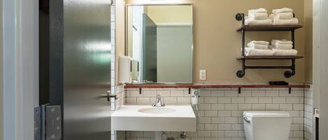 Un baño con un lavabo blanco, un inodoro y un espejo sobre el lavabo. Hay toallas apiladas en un estante sobre el espejo.