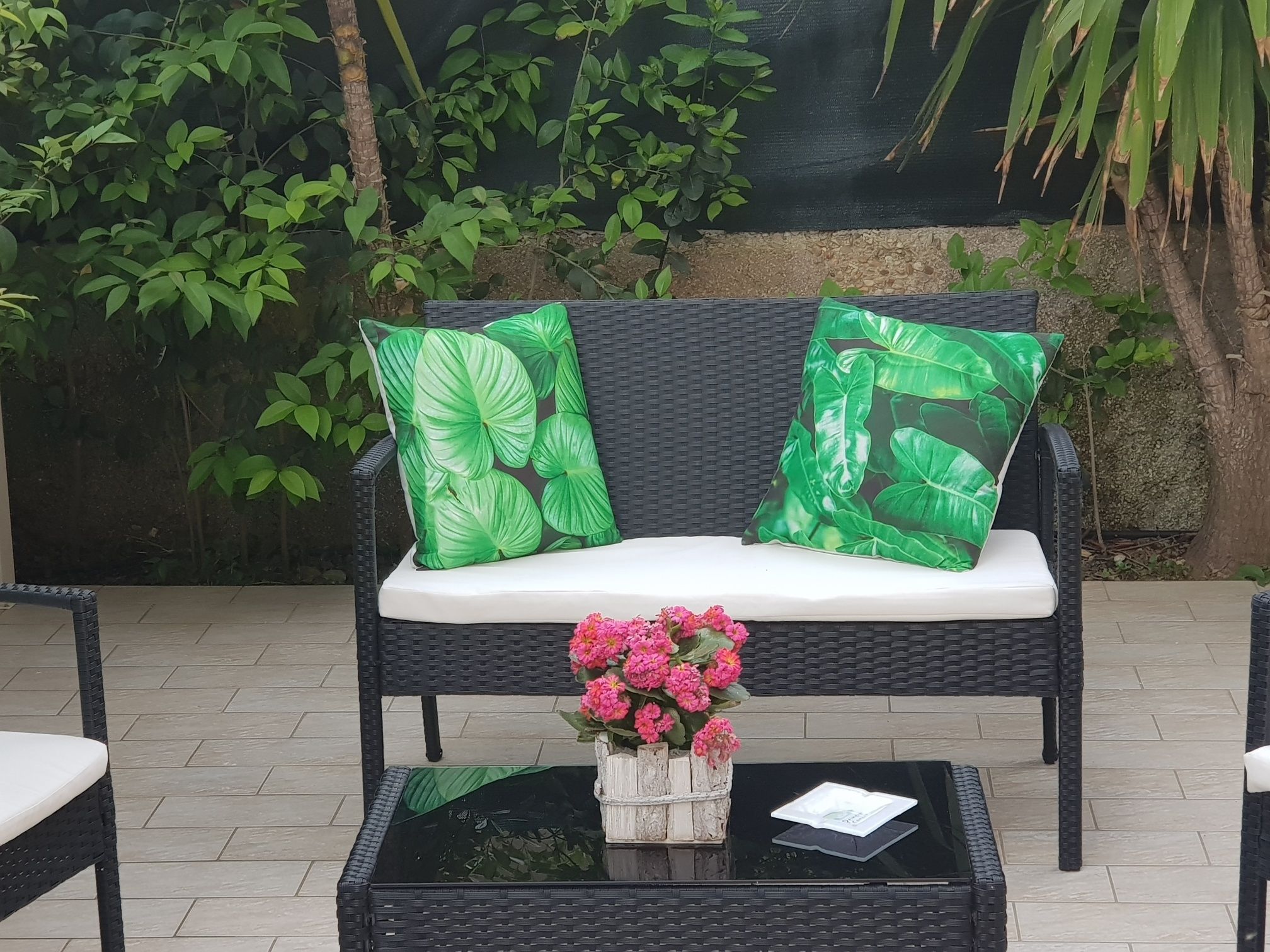 Un sofá de mimbre con cojines de estampado de hojas verdes, una mesita con un jarrón de flores rosas y una bandeja negra.