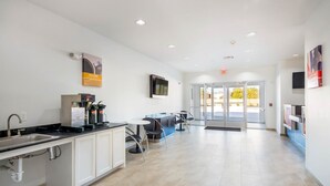 A modern kitchen with a sink, coffee machine, and cabinets. There is a seating area with chairs and a table. The room has white walls and a tiled floor. - Motel 6 Channelview, TX (Channelview)