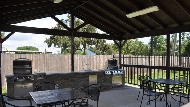 An outdoor patio with a stone barbecue and grill station, metal tables and chairs, and a wooden fence in the background. - Motel 6 Channelview, TX (Channelview)