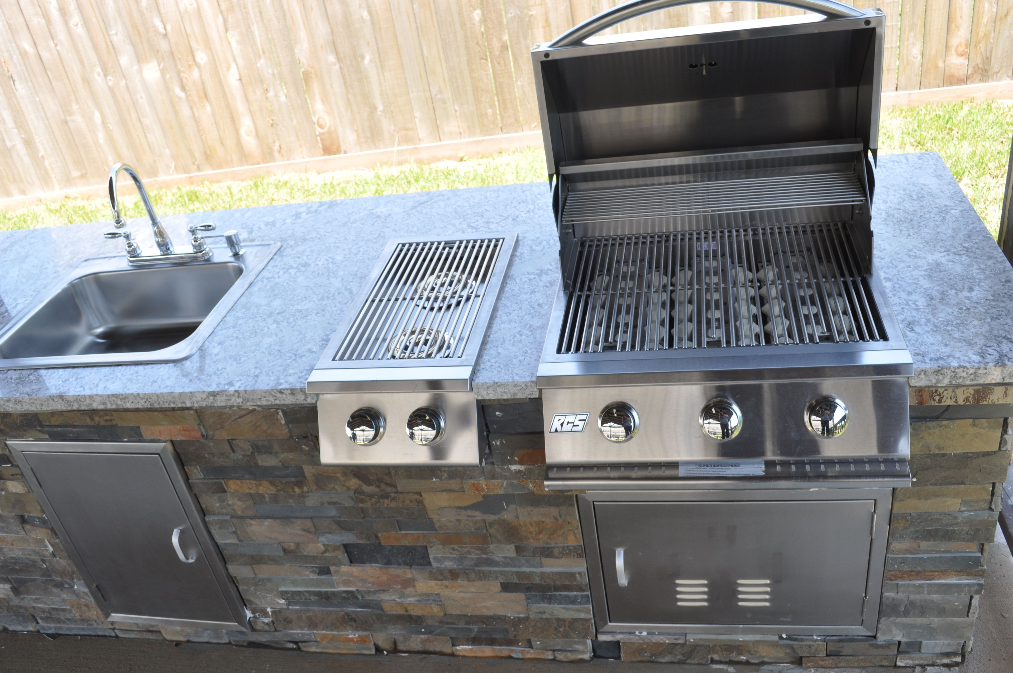 Et udendørs køkken i sten og metal med vask, grill og opbevaringsskabe.