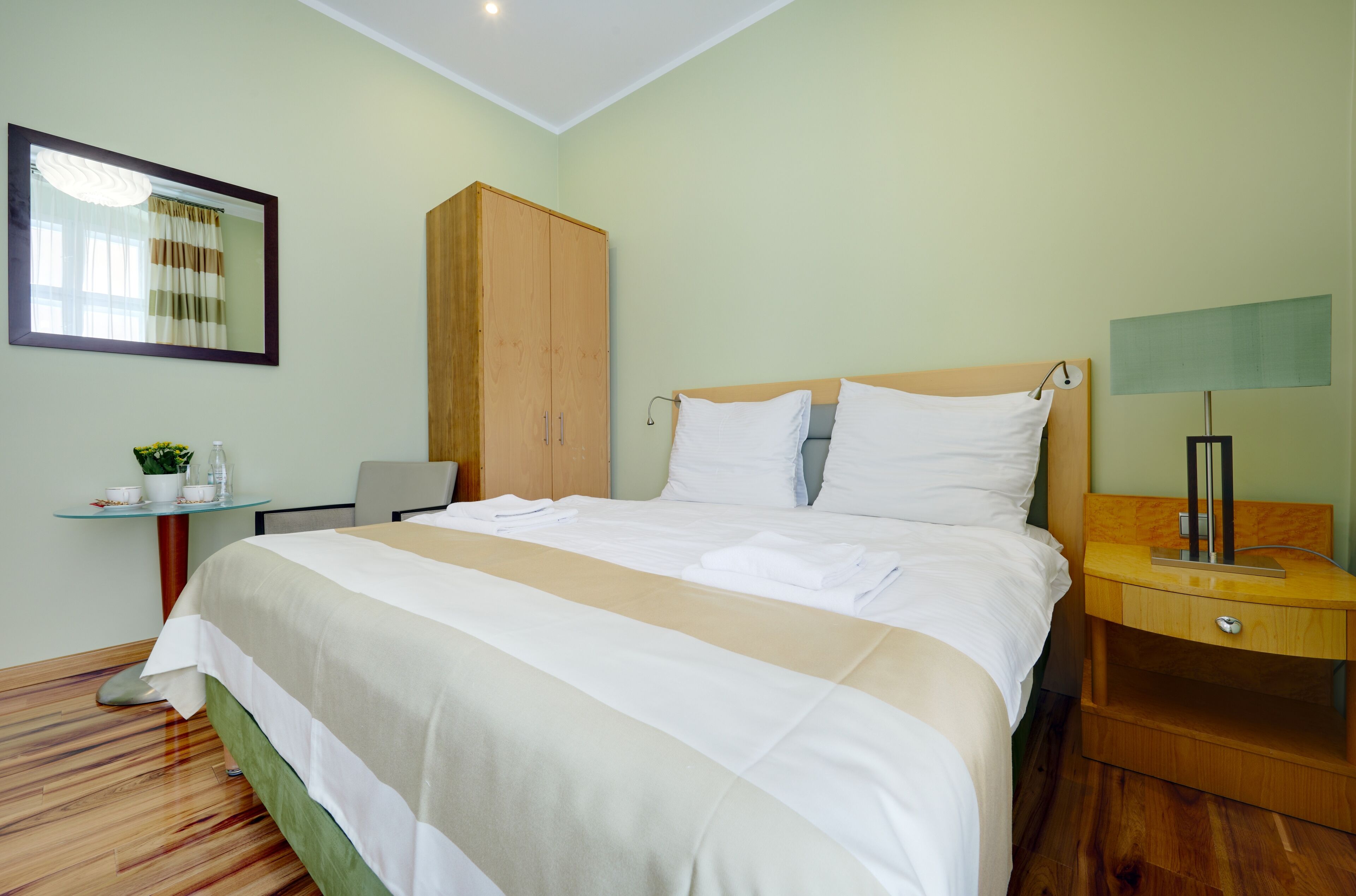 Una cama bien tendida con sábanas blancas, un armario de madera, una mesita con una planta y una lámpara de noche.